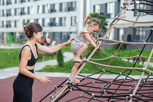 Young mother aids her little daughter as she navigates rope climbing frame at playground with care.