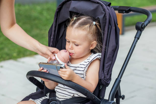 Small child in stroller sips from bottle while curiously looking at smartphon