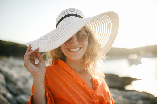 A young woman in a hat standing on a rock by the sea, she is smiling, looking carefree. Small cove at sunset in background