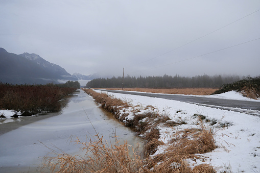 Looking down a ditch along Rannie Road near the Grant Narrows Regional Park and Pitt River Dike during a snowy winter season in Pitt Meadows, British Columbia, Canada.