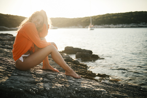 A young woman in bikini and shirt sitting on a rock by the sea, she is smiling, looking carefree. Small cove at sunset in background