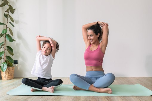 Healthy Latin American mother and her elementary-aged kid stretching their arms on an exercise mat during yoga class together at home for body care with wellbeing and positive emotion.