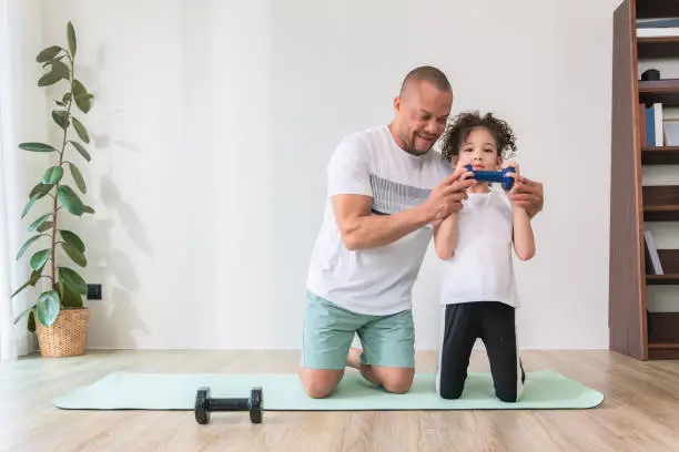 Focused African father training his curly-hair daughter about strength training by using dumbbell in the fitness class at the cozy living room for healthy lifestyle and body care.