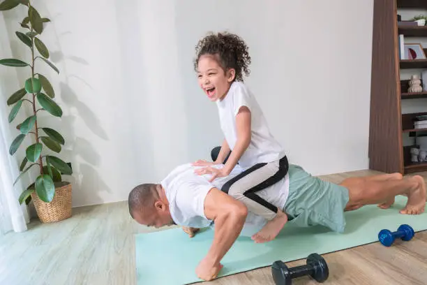 Smiling elementary-aged daughter with curly-hair sitting on a back of her African father while her father training push-up on an exercise mat for increasing his strength at the home gym.