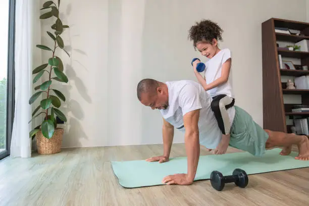 African father with casual clothing exercising by push-up on the exercising mat while his curly-hair daughter sitting on his back and weight training using dumbbell. Happy family exercising together on weekend.