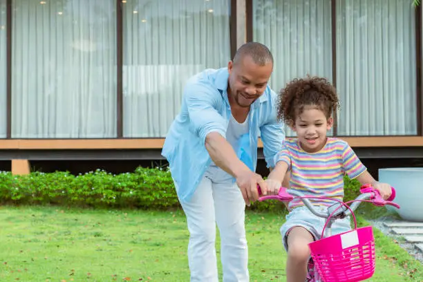 Close up shot of African father spending leisure time on instructing his curly-hair elementary-aged daughter for riding pink bicycle in the yard during weekend for exercising.