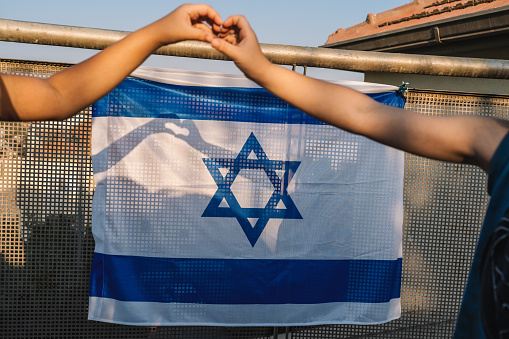 Hands of a Girl and a Boy Form a Heart Shape on the Israeli Flag, with Sunlight Casting a Shadow on the National Flag Hanging from the Balcony.
