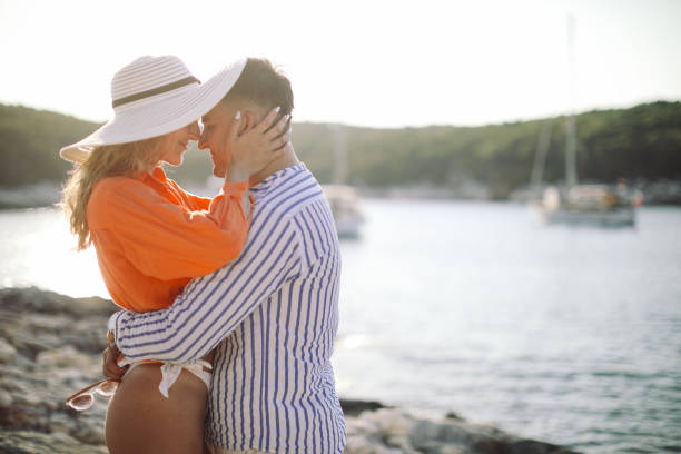 A young couple standing on a rock by the sea, hugging, looking at each other and enjoying sunset. They look happy and carefree.