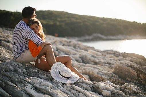 A young couple sitting on a rock by the sea, hugging and enjoying sunset. They look happy and carefree.