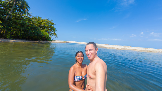 Cheerful couple in swimwear at tropical river edge, clear blue sky.
