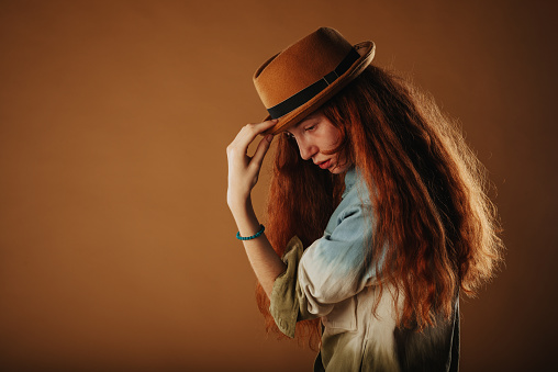 Ginger girl posing with her hand on her hat while standing at the studio
