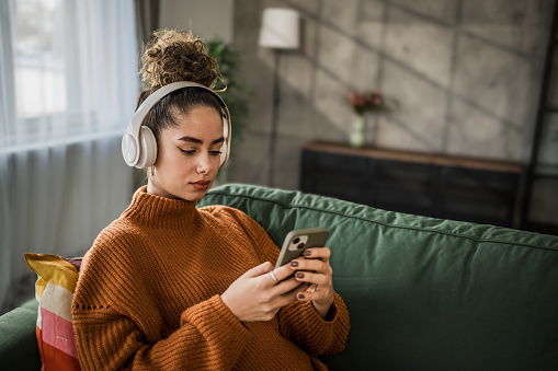 Excited young woman listening to music on wireless headphones over a mobile app while sitting on a comfortable sofa