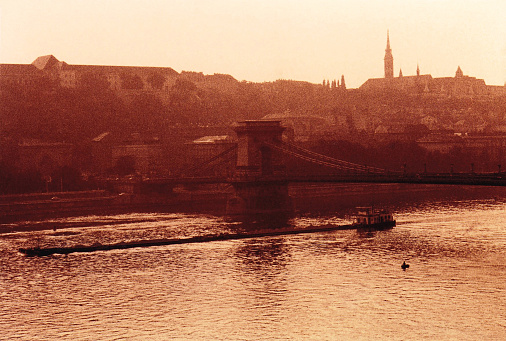 Budapest emerges in timeless sepia tones. The Chain Bridge gracefully spans the Danube as a flat ship glides beneath, laden with sand. On the opposite bank, historic structures stand against wavy river currents, crafting an enduring and captivating tableau.