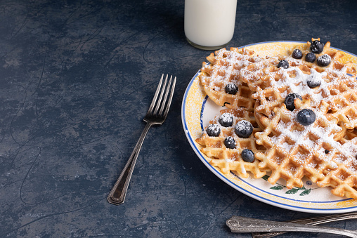 Waffles, blueberries, and powdered sugar on a blue tabletop with a glass of milk and linen napkin.