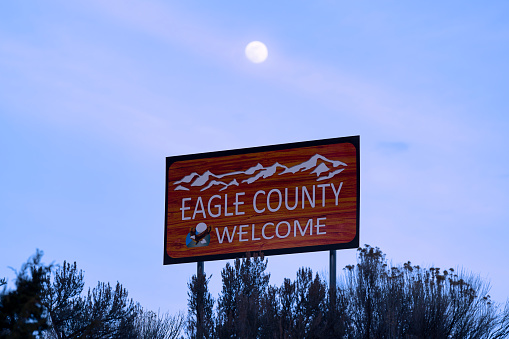 Welcome to Eagle County Road Sign - Sign along Interste 70 at county line from Garfield into Eagle County, Colorado USA. Large sign with mountains against dusk blue sky and moon in sky.