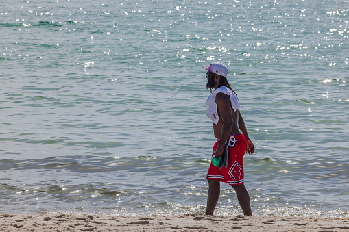 USA. Miami Beach. 01.24.2024. A handsome young African-American man with an athletic build, wearing red swimming shorts and a white cap, strolling along the Miami Beach shore. USA.