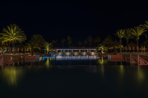 Willemstad. Curacao. 01.25.2024. A beautiful night view of the hotel grounds with an open pool, a bar, and palm trees reflecting in the water.