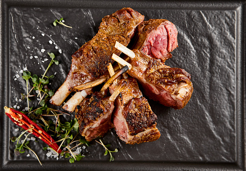 Juicy grilled lamb rack with a crusty sear, accented by fresh green herbs and a fiery red chili on a dark slate plate.