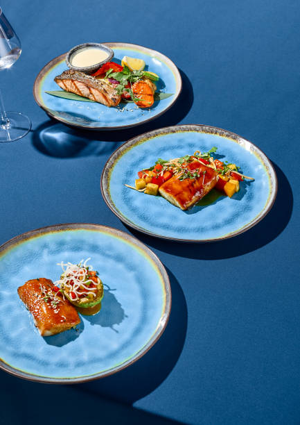 A set of Asian-style dishes featuring salmon, cod, and pike perch, presented on blue ceramic plates against a monochromatic blue background with sharp shadows