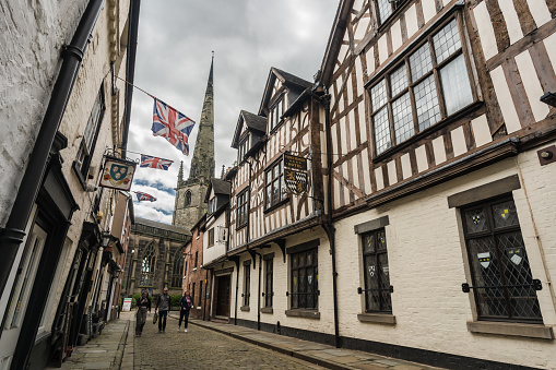 Shrewsbury, Shropshire, England, May 1st 2023. People walking down cobbled Tudor alley with church in background, editorial travel illustration.