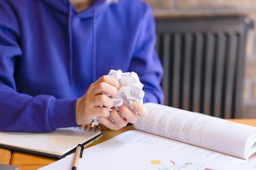 Crumpled sheet of paper in hands close-up, girl in blue sweater. Photo of female hands crumpling paper, ruined note, paper garbage, textbook on wooden table, pen and notebook