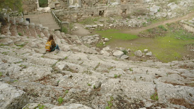 A female tourist with a backpack descends the steps of the ancient Greek amphitheater and sits at the bottom to admire the view of the ancient structures.