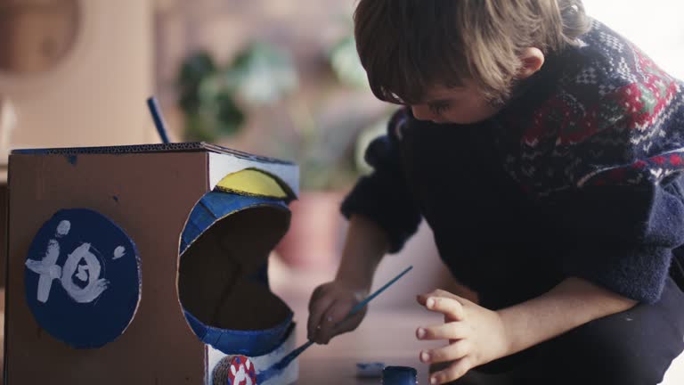 Little child boy painting his space helmet mad of cardboard.