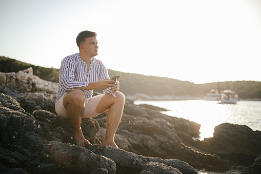 A young man in shirt and shorts sitting on a rock by the sea, enjoying sunset. He is holding mobile phone in his hands.