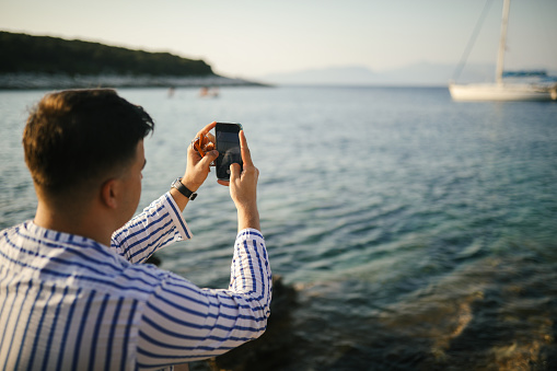 A young man in shirt and shorts sitting on a rock by the sea, enjoying sunset. He is taking photos with mobile phone.