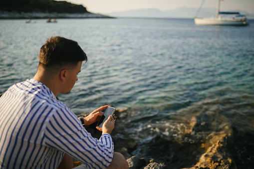 A young man in shirt and shorts sitting on a rock by the sea, enjoying sunset. He is holding mobile phone in his hands.