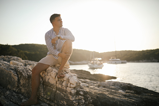 A young man in shirt and shorts sitting on a rock by the sea, enjoying sunset.