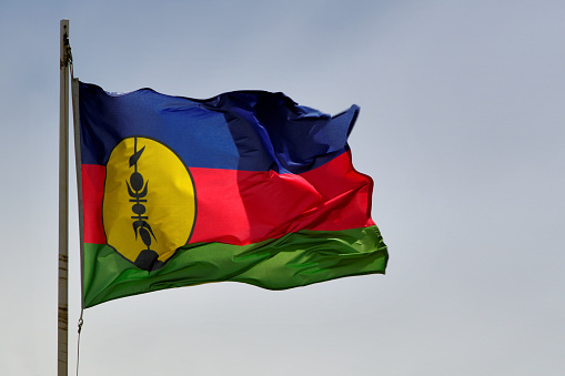 Nouméa, South Province, New Caledonia: Kanak flag / Flag of New Caledonia, indigenous Melanesian tricolor used in parallel with the French flag, originally the flag of the independence movement FLNKS. The Flag consists of three equal horizontal bands of blue (top), red, and green. A large yellow disk edged in black and displays a black 'fleche faitiére' (rooftop arrow). Emblem of the Kanak chiefdom, the 'fleche faitiére', is a traditional element of Kanak housing, it is a wood sculpture which dominates and adorns the cone-shaped thatched roof of the large huts and ceremonial huts of a clan.