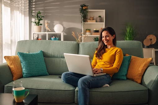 Smiling caucasian woman using laptop while sitting on a mint couch at home. Happy female shopping or chatting online in social network, typing blog, freelancer working on a project.