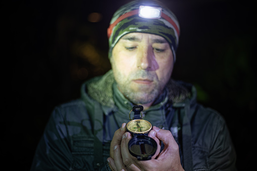 Close-up photo of a traveling man looking at compass in his hand at night. A man uses a headlamp to light the path and check his compass during his nighttime hiking adventure.