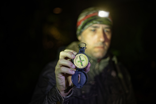 Close-up photo of a traveling man looking at compass in his hand at night. A man uses a headlamp to light the path and check his compass during his nighttime hiking adventure.