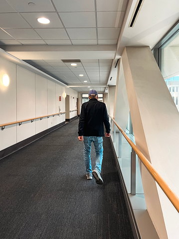 Rear View of a Mature Senior Man Walking Down a Clean, Brightly Lit Hallway. He is dressed casually in jeans and a baseball cap. Modern interior architecture.
