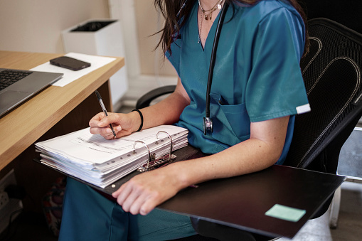 Close-up of a female doctor wearing scrubs writing notes in a binder while working at her office desk in a hospital