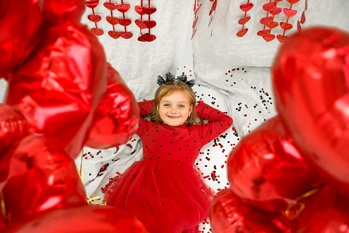 A little girl in a red polka dot dress, surrounded by heart-shaped balloons, lies on the floor looking at the camera.