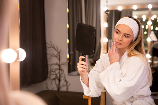 Young, blonde, and beautiful, a woman in her mid-20s sits in front of a large mirror with lights, wearing a white bathrobe. In her beige and brown modern room, she applies makeup, enhancing her stunning features. This stock image captures the elegance of a refined beauty routine, ideal for projects highlighting the artistry of makeup and modern aesthetics.
