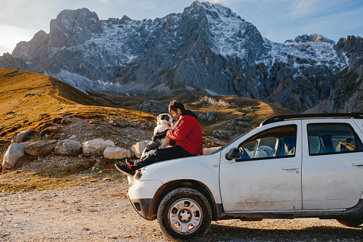 A young man sitting on the hood of his 4wd SUV car with his border collie dog on a break while driving on a mountain adventure trip.