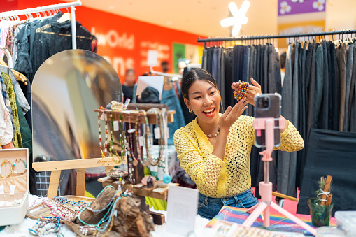 Asian woman using smartphone live streaming presentation fashion handmade jewelry selling online shop during open jewelry shop kiosk at shopping mall. Small business owner and handmade concept.