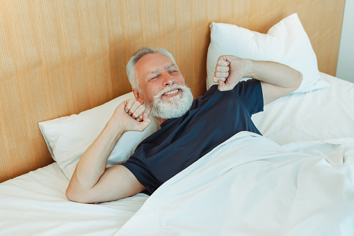 Portrait of happy bearded senior man stretches, wake up lying on comfortable bed. Morning concept