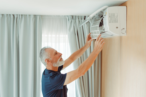 Portrait of attractive senior serviceman checking air conditioner, replacing, cleaning air conditioner filter in apartment. Elderly worker repairing equipment. Concept technology