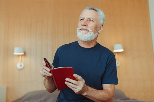 Attractive positive senior man rhyming book while sitting on bed in hotel, looking away. Handsome gray haired grandfather reading. Education concept