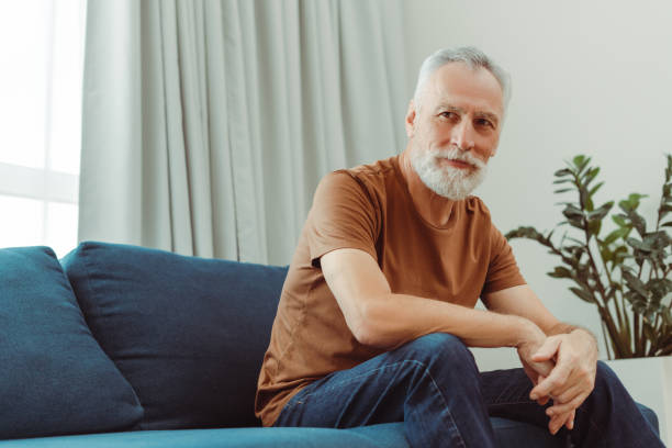Elderly man smiling sitting on soft sofa in living room and looking away. Advertisement concept