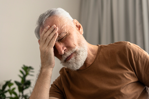 Portrait of tired senior man with closed eyes touching head, having headache, migraine in living room. Sad pensioner will receive sad news. Treatment concept