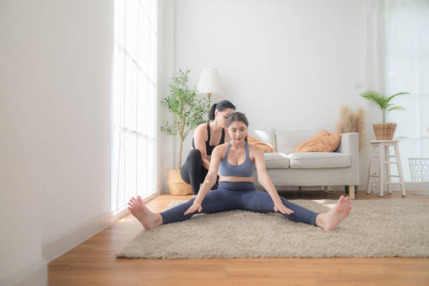 two women confident training yoga. athletic women in sportswear doing fitness stretching exercises at home in the living room. sport and recreation concept. yoga teacher is helping young woman. - yoga business women living room ストックフォトと画像