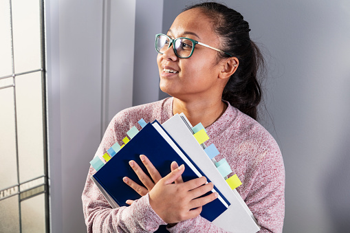 A young Asian woman, a university student in her 20s, holding two textbooks to her chest. She has used green, blue, and yellow page markers to color-code information for reference.
