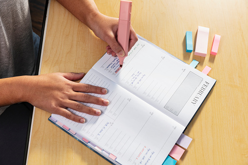 Hands of a young Asian woman, a university student, highlighting an item in her personal organizer. She is color coding her book with pink, purple and blue page markers.