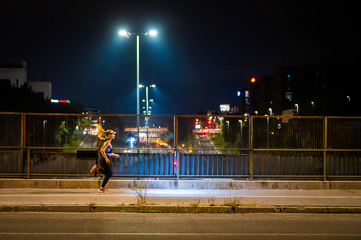 Woman jogging at night, crossing urban bridge illuminated with street lights, wide shot side view